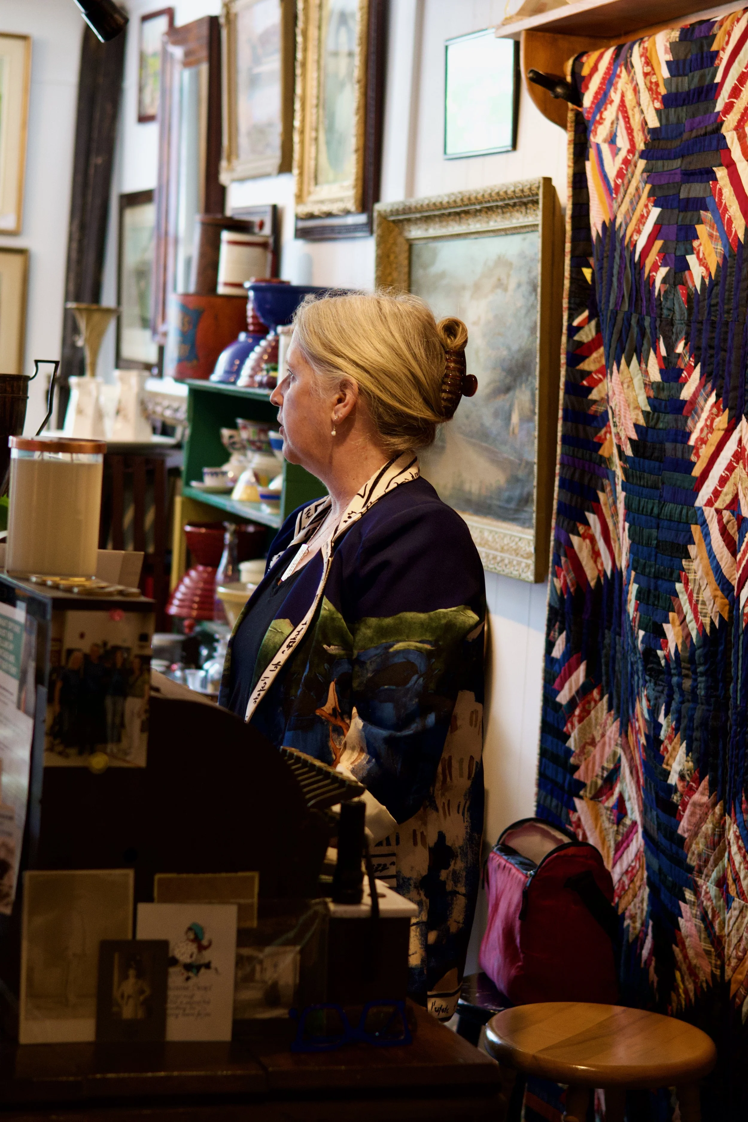 A woman with blonde hair styled in a bun is standing in a vintage shop or gallery, surrounded by framed artwork on the walls and various decorative items on shelves. She is wearing a dark blazer with a patterned shirt underneath. In the foreground, there are postcards or greeting cards on display, and to her right, a colorful quilt with a geometric mosaic pattern hangs on the wall. A round wooden stool is visible nearby, and the space appears cozy and eclectic.