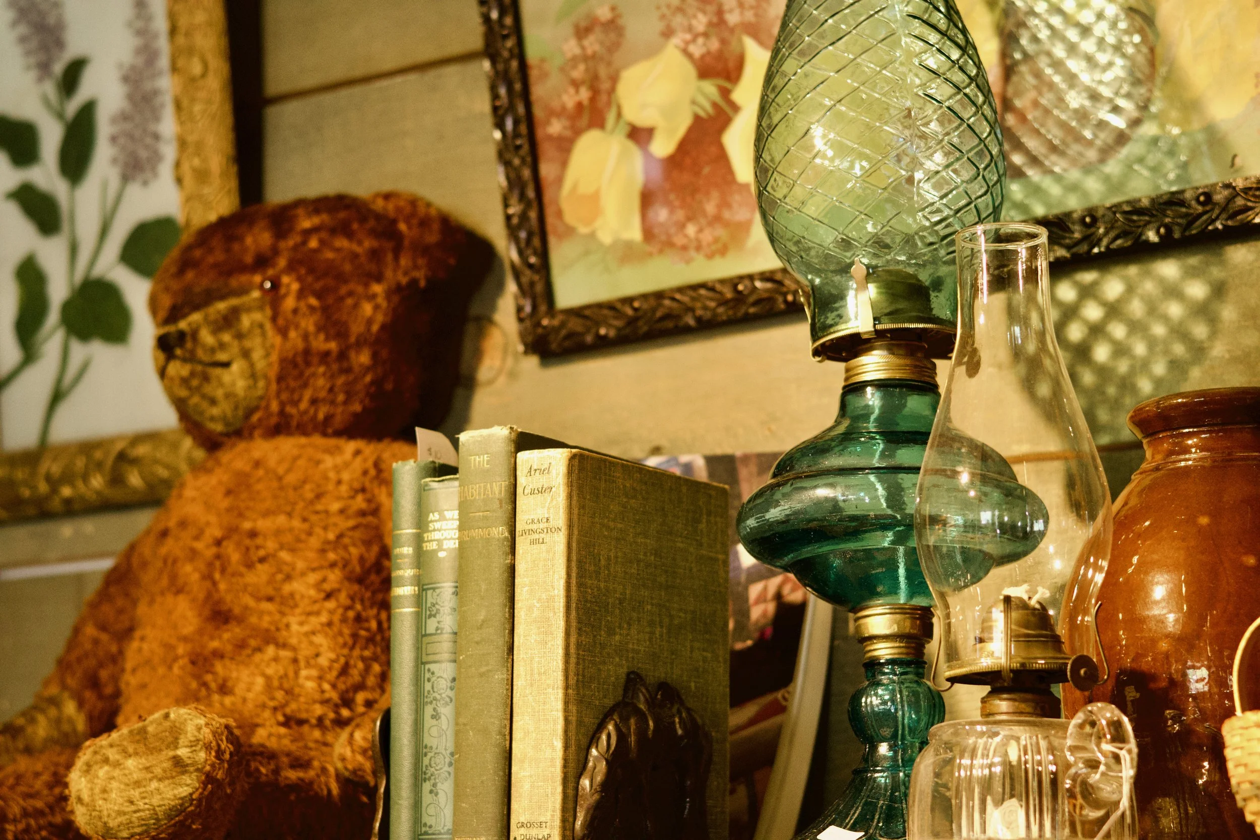 A decorative shelf displaying an assortment of glass vases, a framed floral artwork, a couple of books, and ceramic items, with a leafy patterned wall in the background.