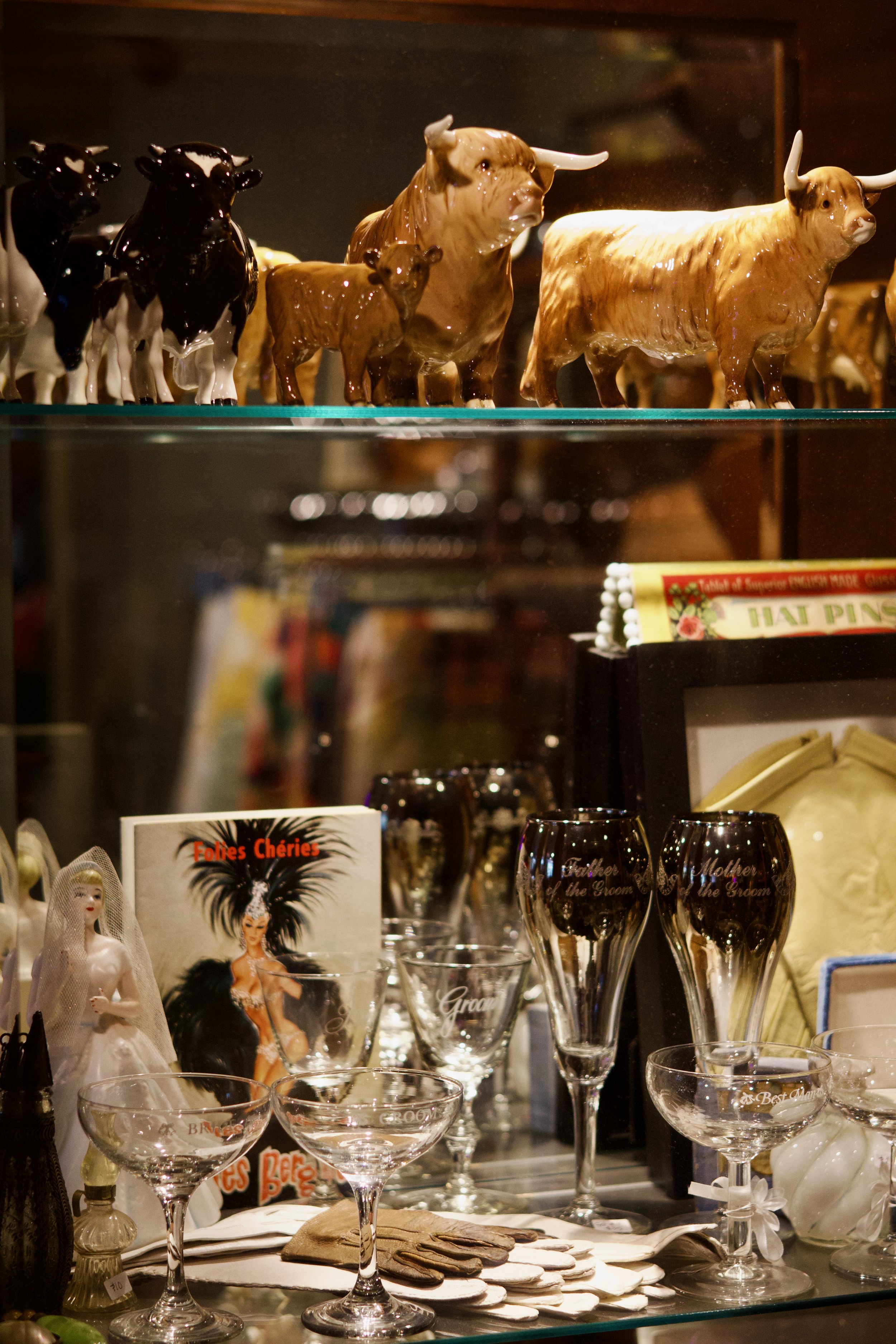 A display of various decorative ceramic figurines and glassware on shelves. The figurines include black and brown cows and realistic bulls. The glassware includes clear glasses with inscriptions and champagne flutes, some of which have inscriptions like 'Father of the Groom' and 'Mother of the Groom'. There are also dolls, books, and miscellaneous items on the lower shelf.