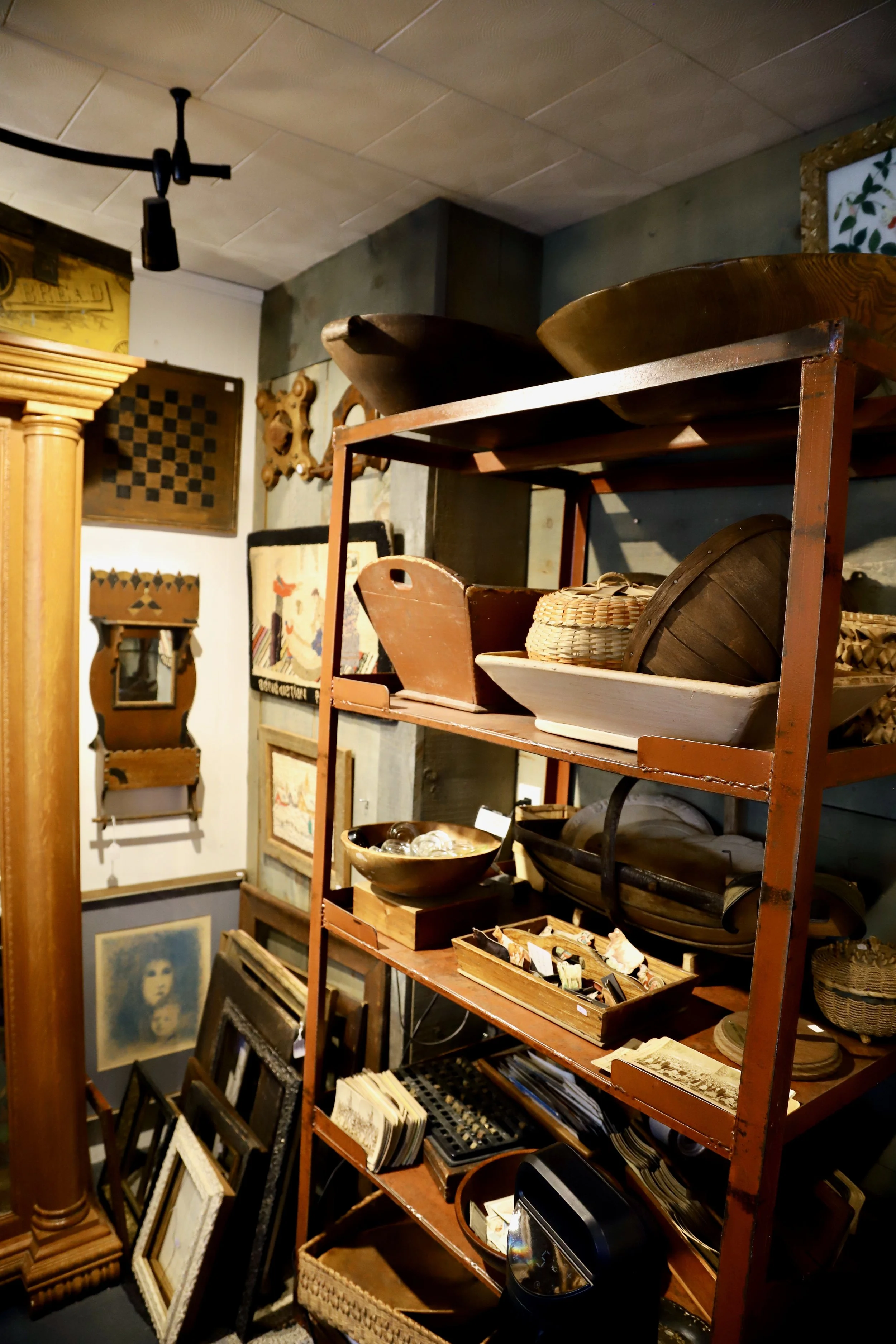 A wooden shelf containing various vintage items such as bowls, baskets, and picture frames, with artwork and decor on the wall in the background.