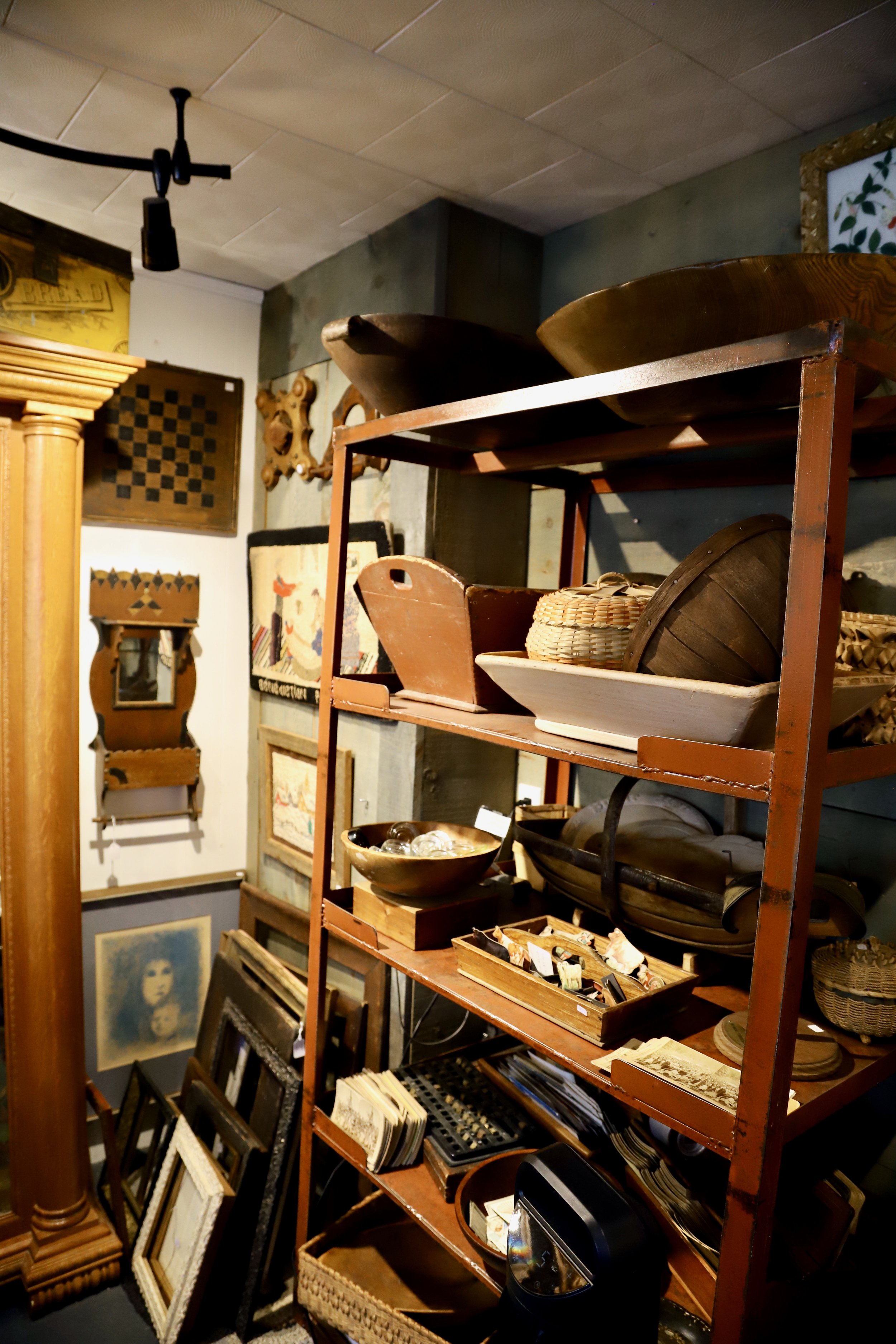 A tall red metal shelf displaying various vintage items like bowls, baskets, and trays, with framed artwork and mirrors on the wall behind it, in an antique store.