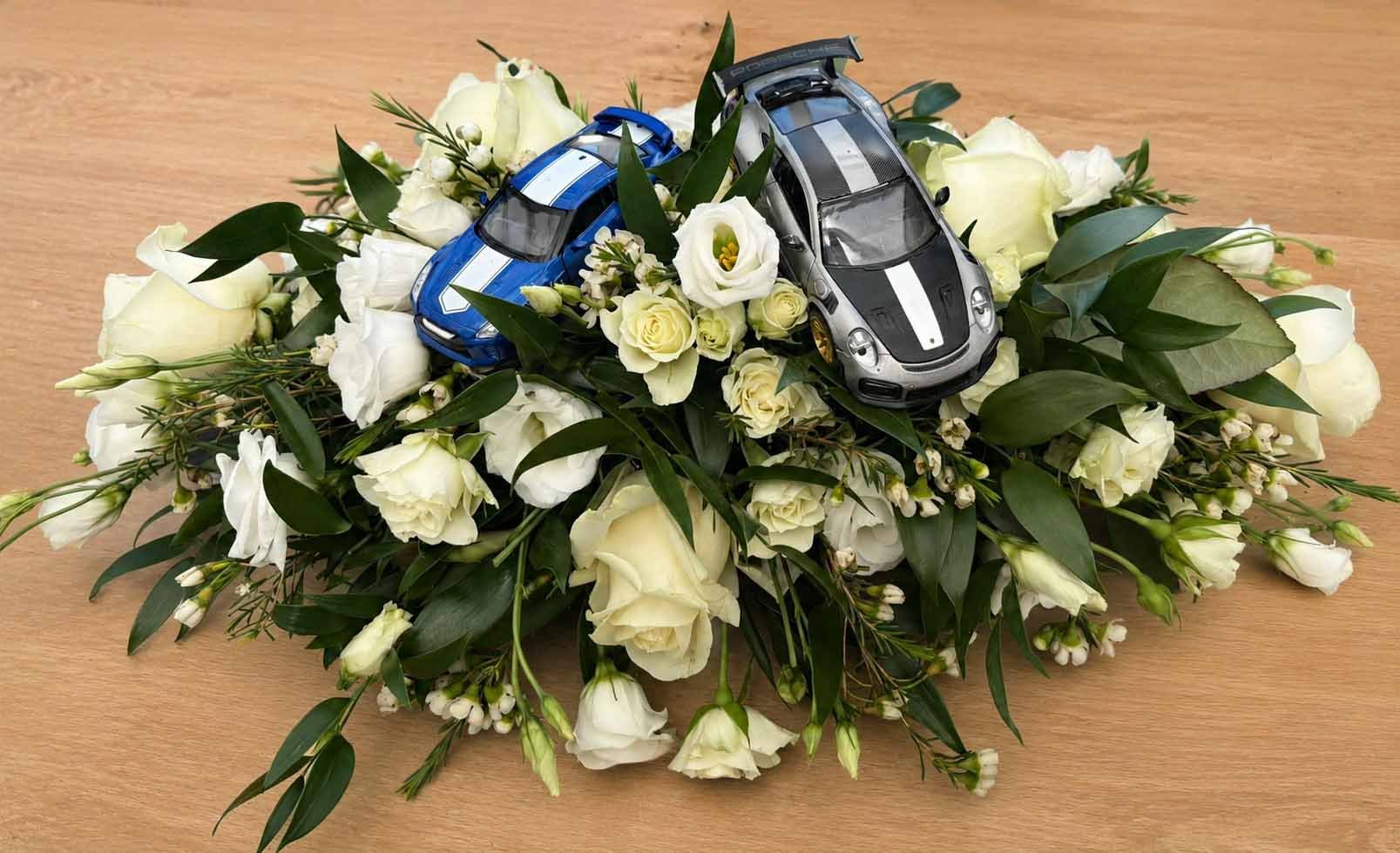 Two toy cars, one blue and one black with white racing stripes, placed on a bed of white flowers and green leaves on a wooden surface.