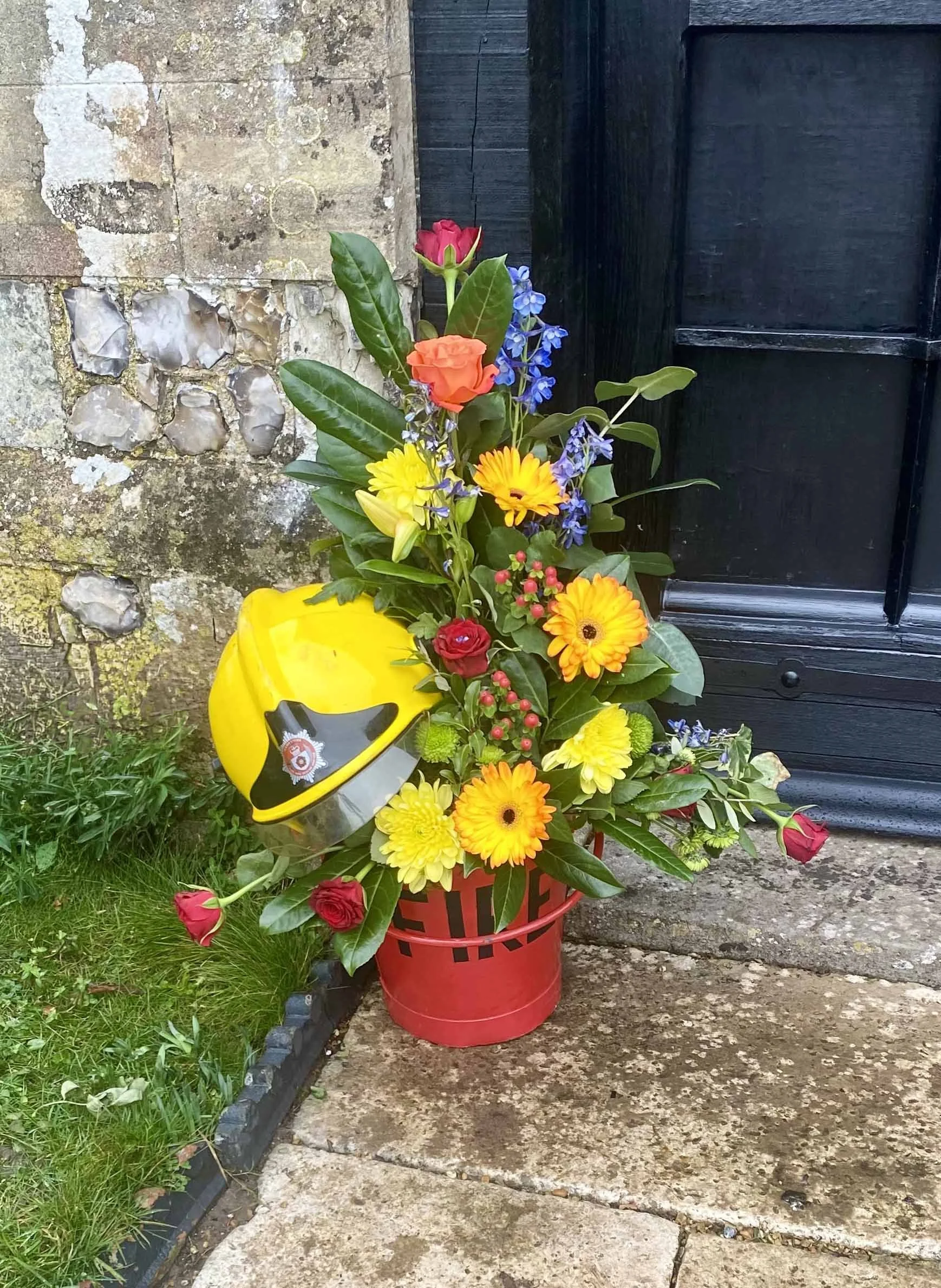 A flower arrangement with various colorful flowers, including red roses, yellow daisies, and purple bluebells, placed in a red basket with a firefighter's yellow helmet on top, outside near a stone wall and a black wooden door.