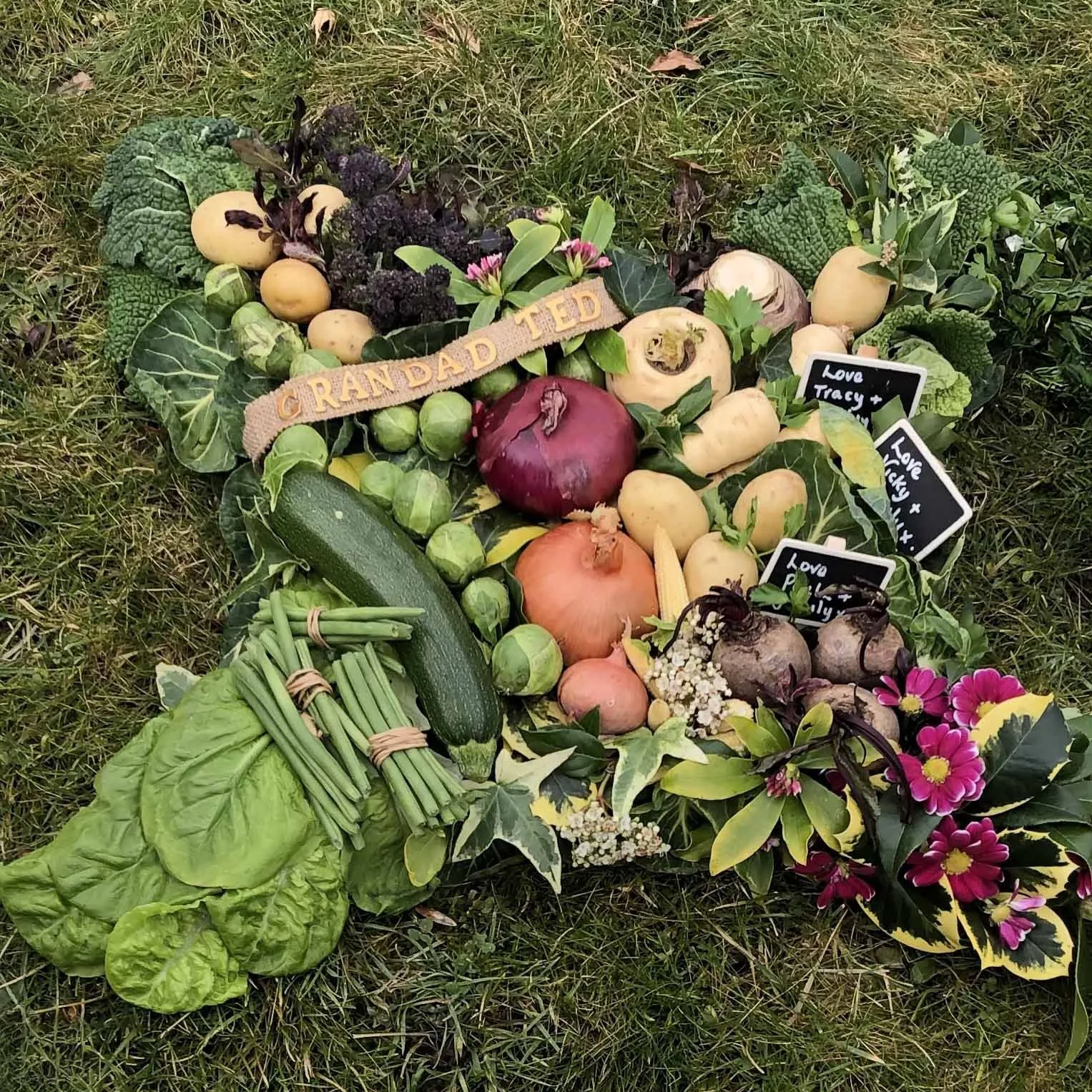 A collection of various fresh vegetables arranged on grass, including lettuce, zucchinis, brussels sprouts, onions, beets, and decorative flowers, with small signs that say 'Love Tracy' and 'Love Picky'.