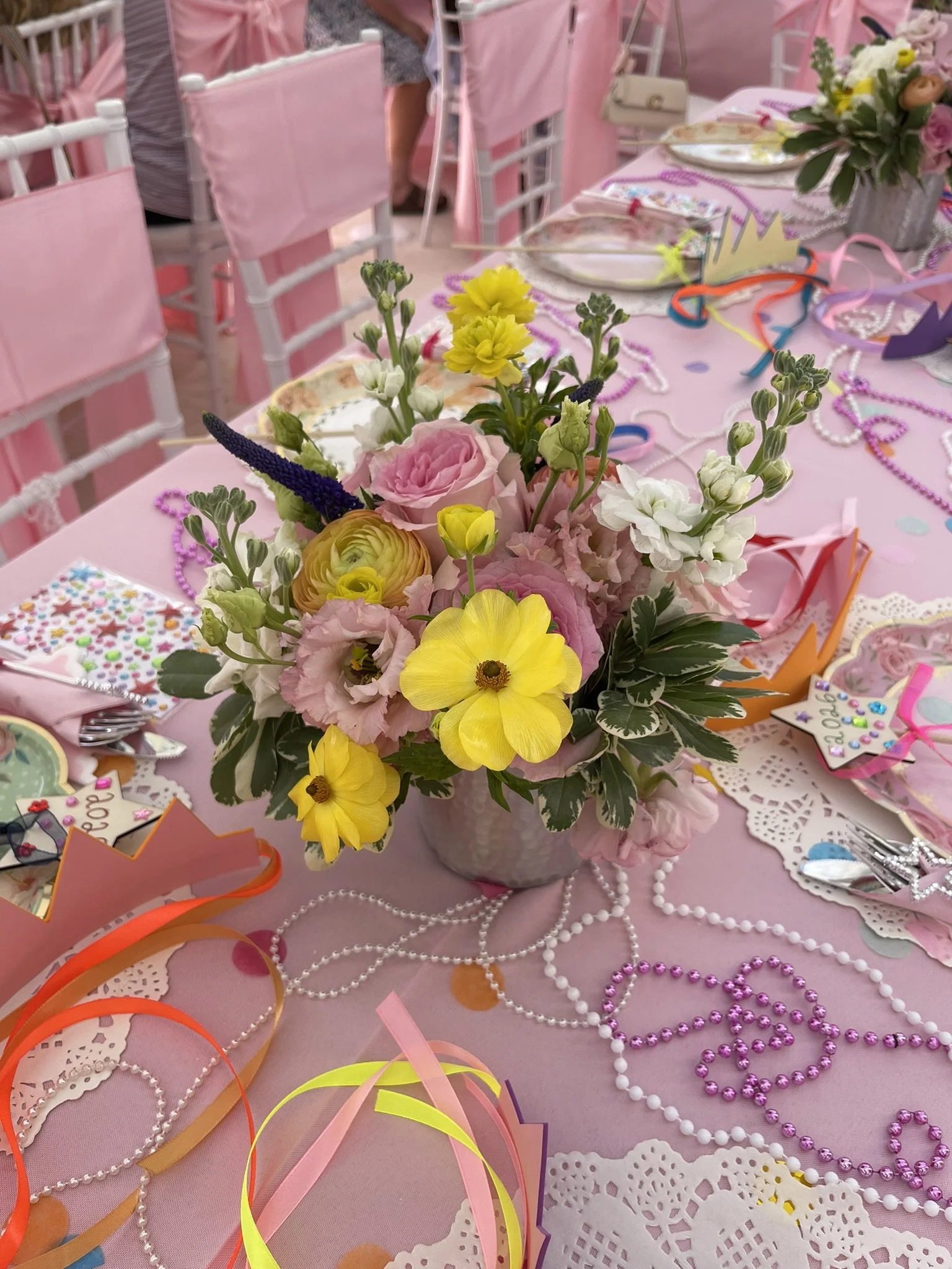 A pink table decorated for a celebration with a colorful floral centerpiece, pearl necklaces, paper cutouts, and party accessories including ribbons and decorative plates.