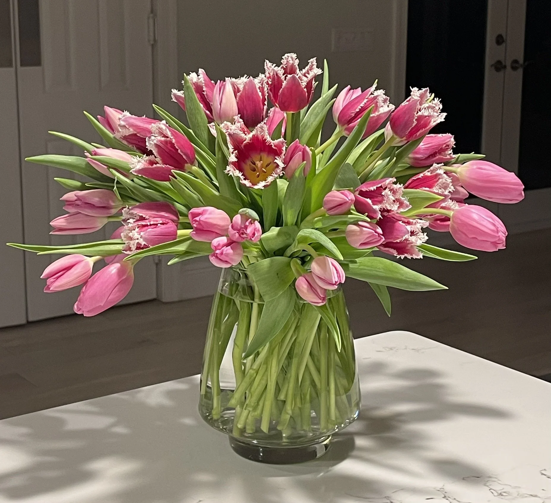 A clear glass vase filled with pink tulips and green leaves sitting on a white surface.