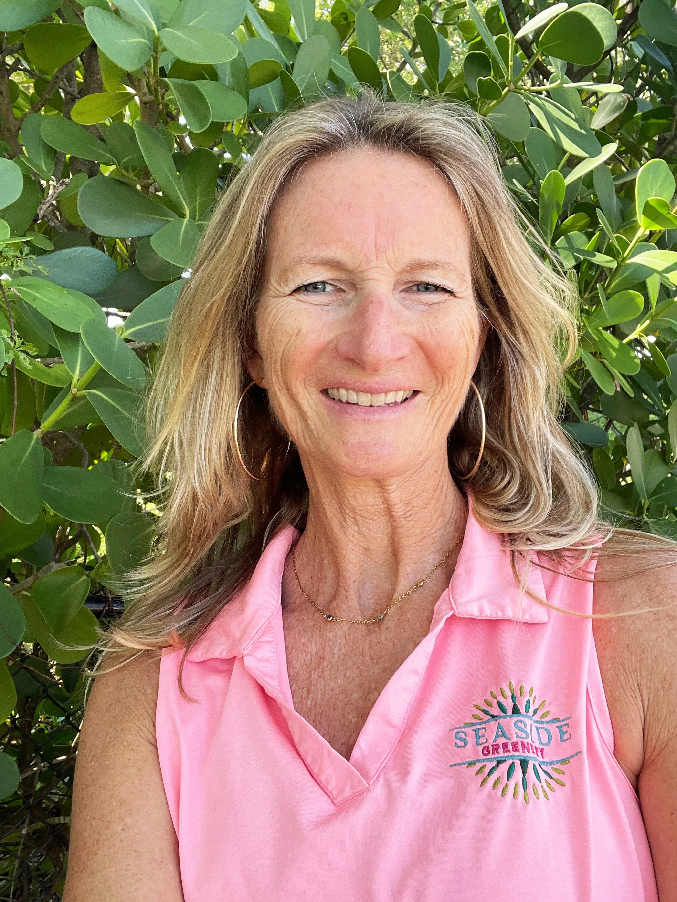A smiling woman with blonde hair and blue eyes, wearing hoop earrings, a gold necklace, and a pink sleeveless shirt with a logo that reads 'SEASIDE GREENWAY,' standing in front of green leafy bushes.