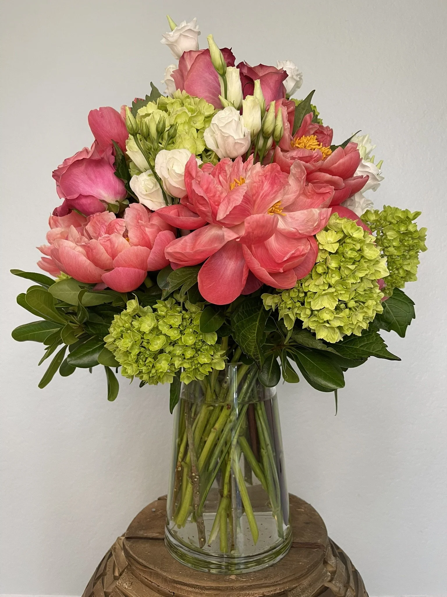 A bouquet of colorful flowers including pink roses, white lisianthus, green hydrangeas, and other mixed blooms in a clear glass vase on a wooden surface against a white wall.