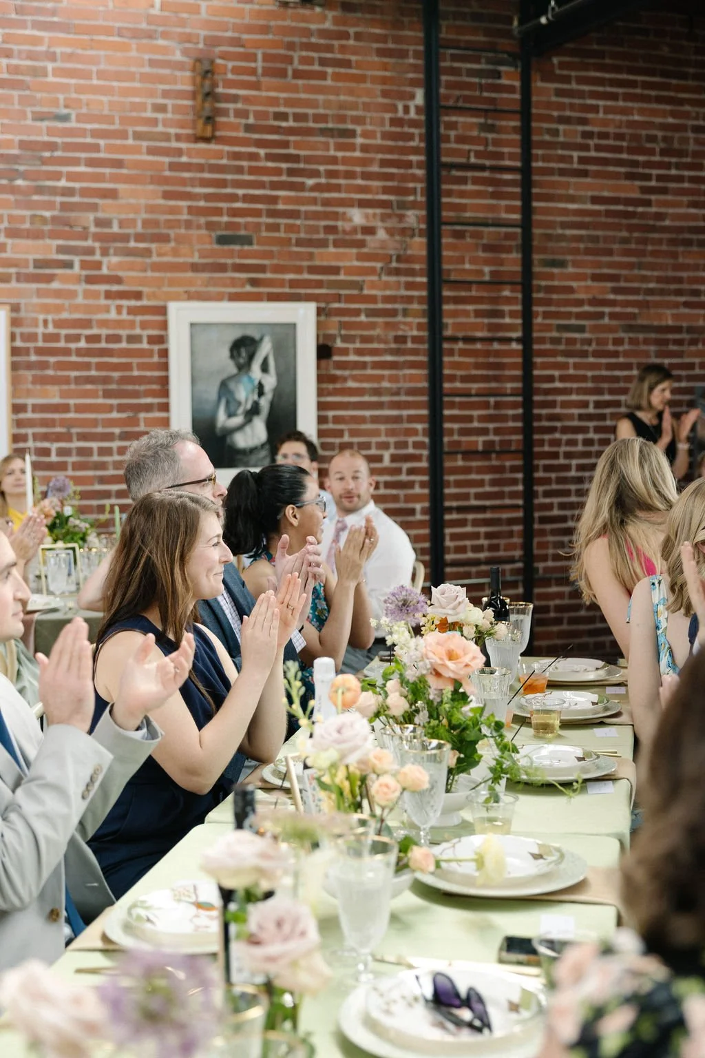 People seated at a banquet table, clapping, with a brick wall and framed black-and-white photograph of a woman in the background.