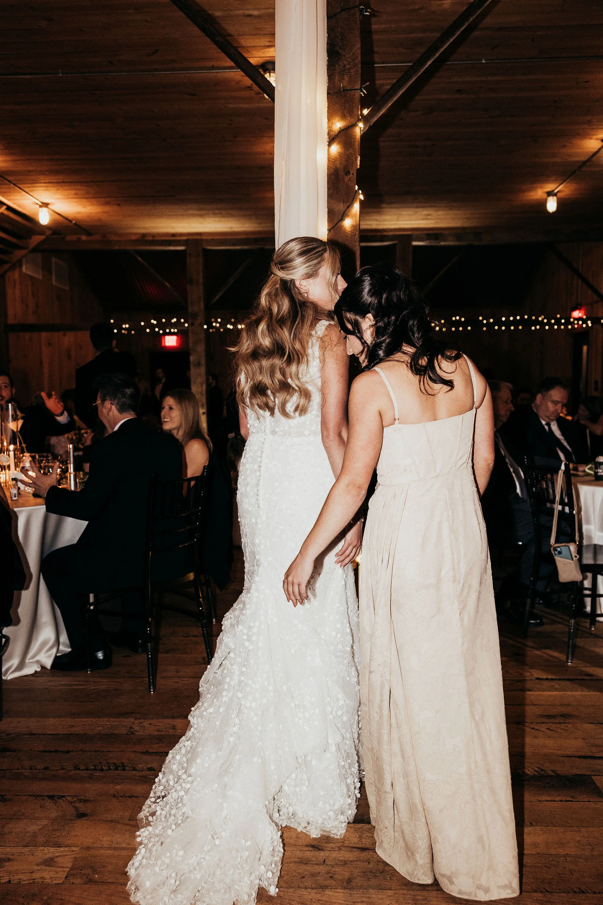 Two women dressed in wedding gowns sharing a tender moment at a reception, with guests seated at tables in the background. The setting has a rustic, wooden decor with string lights overhead.