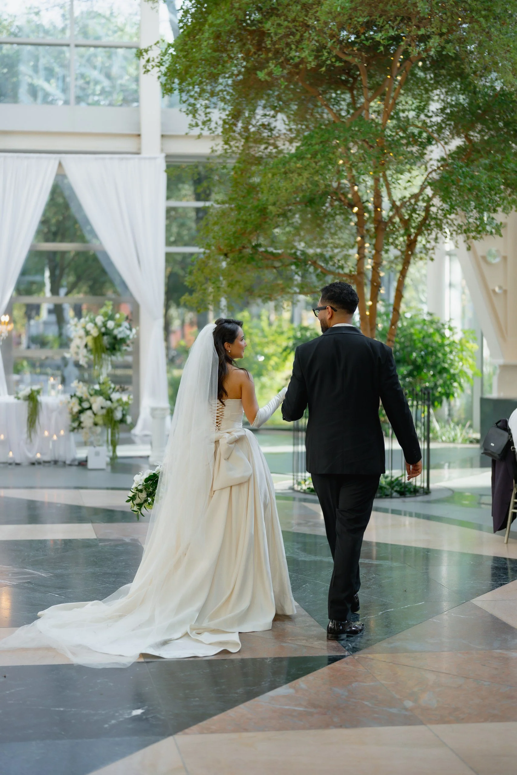 A bride and groom holding hands during their wedding ceremony in a bright, modern indoor venue with large windows, white curtains, and floral arrangements.