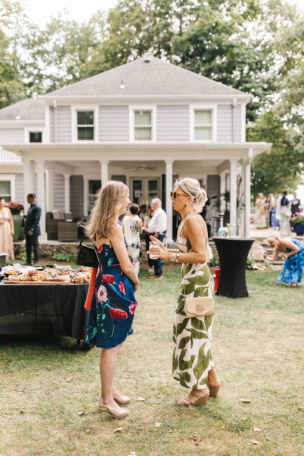 Two women are talking and smiling at an outdoor gathering or party in a backyard, with a large house in the background. One woman has blonde hair and is wearing sunglasses and a patterned dress, while the other woman has light brown hair and is weari