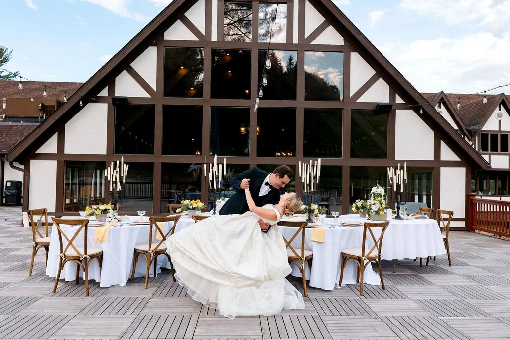 A newlywed couple dancing outside on a deck with decorated tables for a wedding reception, in front of a large rustic-style building with large glass windows.