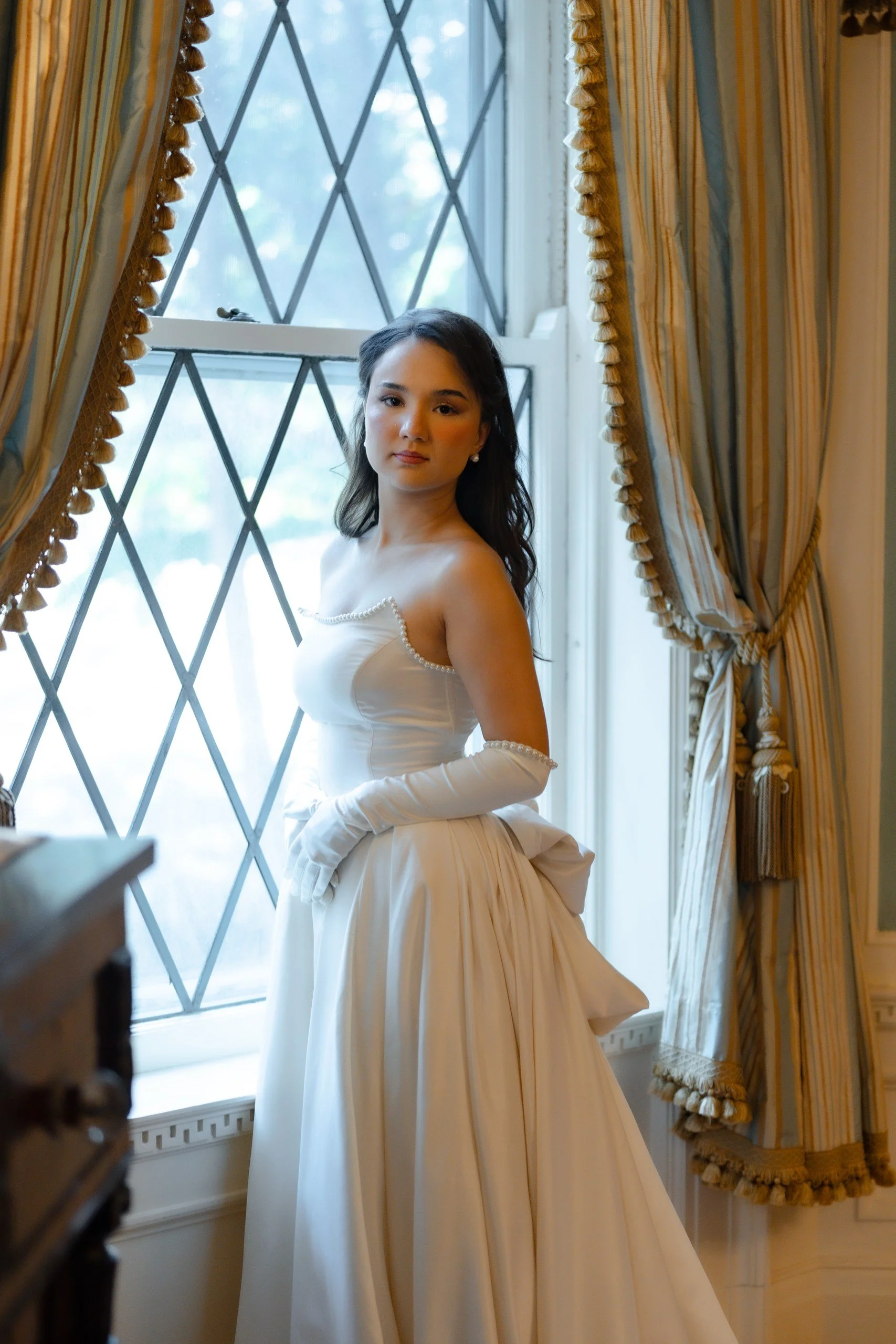 A woman with long dark hair wearing a white Victorian-style dress with pearl details on the shoulders, standing near a window with ornate gold and striped curtains, inside a room with classic decor.