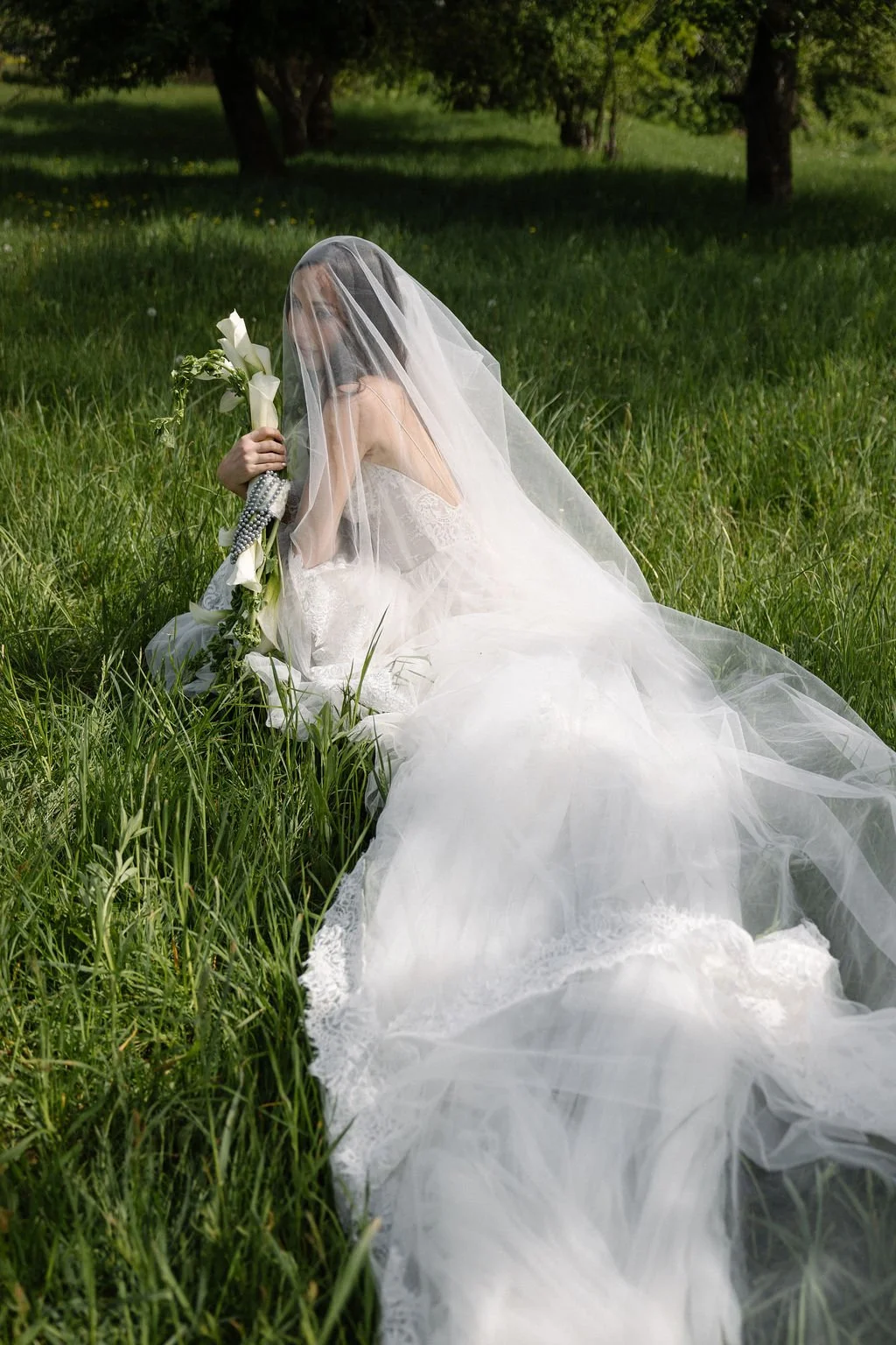 A woman in a wedding dress with a veil sitting on grass in a field, holding a bouquet of white flowers.