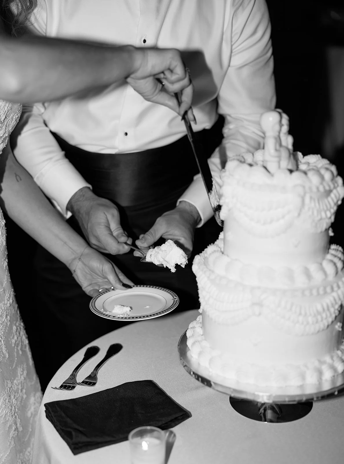Black and white photo of a wedding cake being sliced by a couple at their wedding reception, with part of the cake visible and a decorated tiered cake in the foreground.