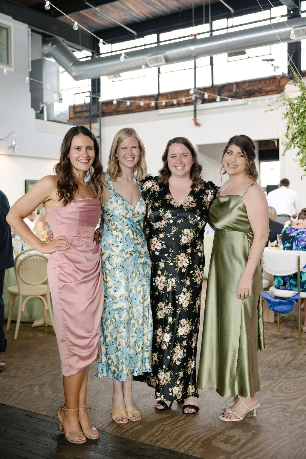 Four women in floral and satin dresses standing together indoors at a social event, smiling at the camera.