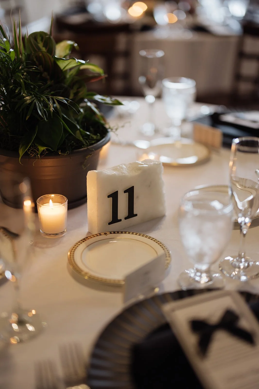 A formal dining table with a large potted plant, a lit candle in a glass holder, a white table number block with the number 11, various wine glasses, and a set plate with gold trim, all under warm lighting.