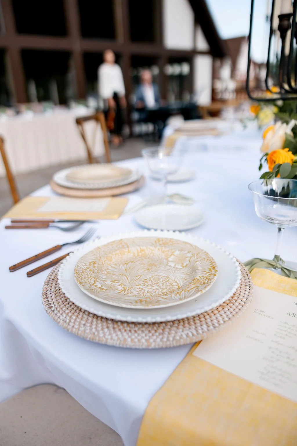 Close-up of a dining table set with decorative plates, cutlery, glasses, a menu, and floral centerpiece, outdoors with people in the background.