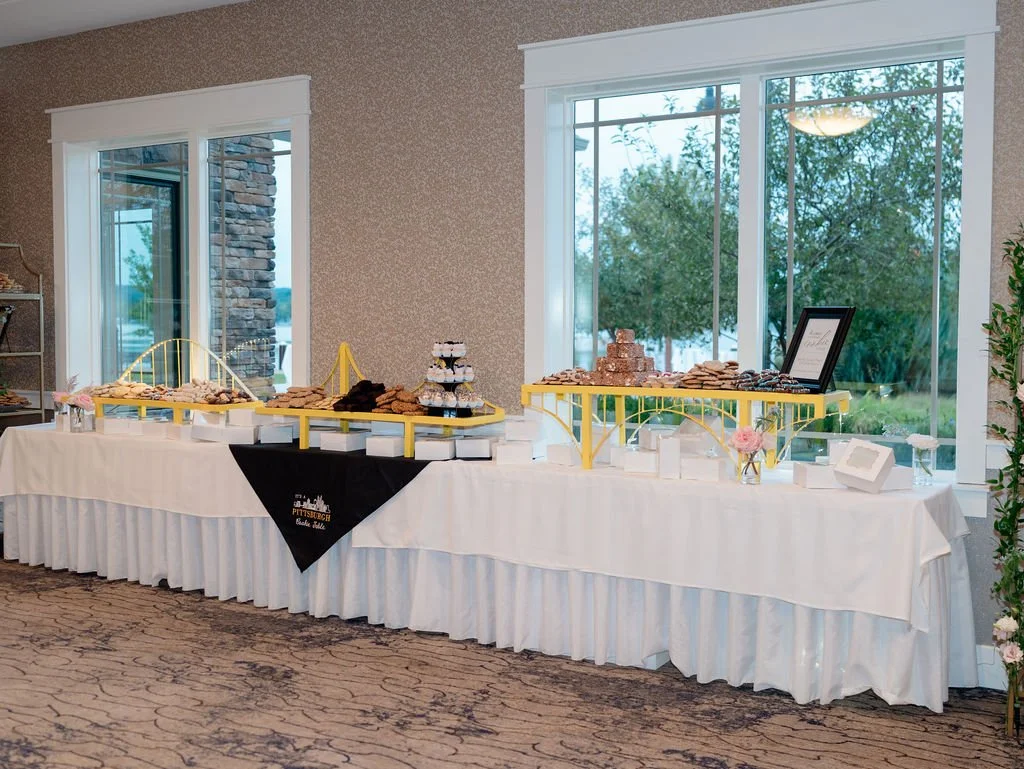A dessert table with various cookies, cupcakes, and other treats, decorated with gold and yellow accents, set up indoors near large windows with a view of trees outside.