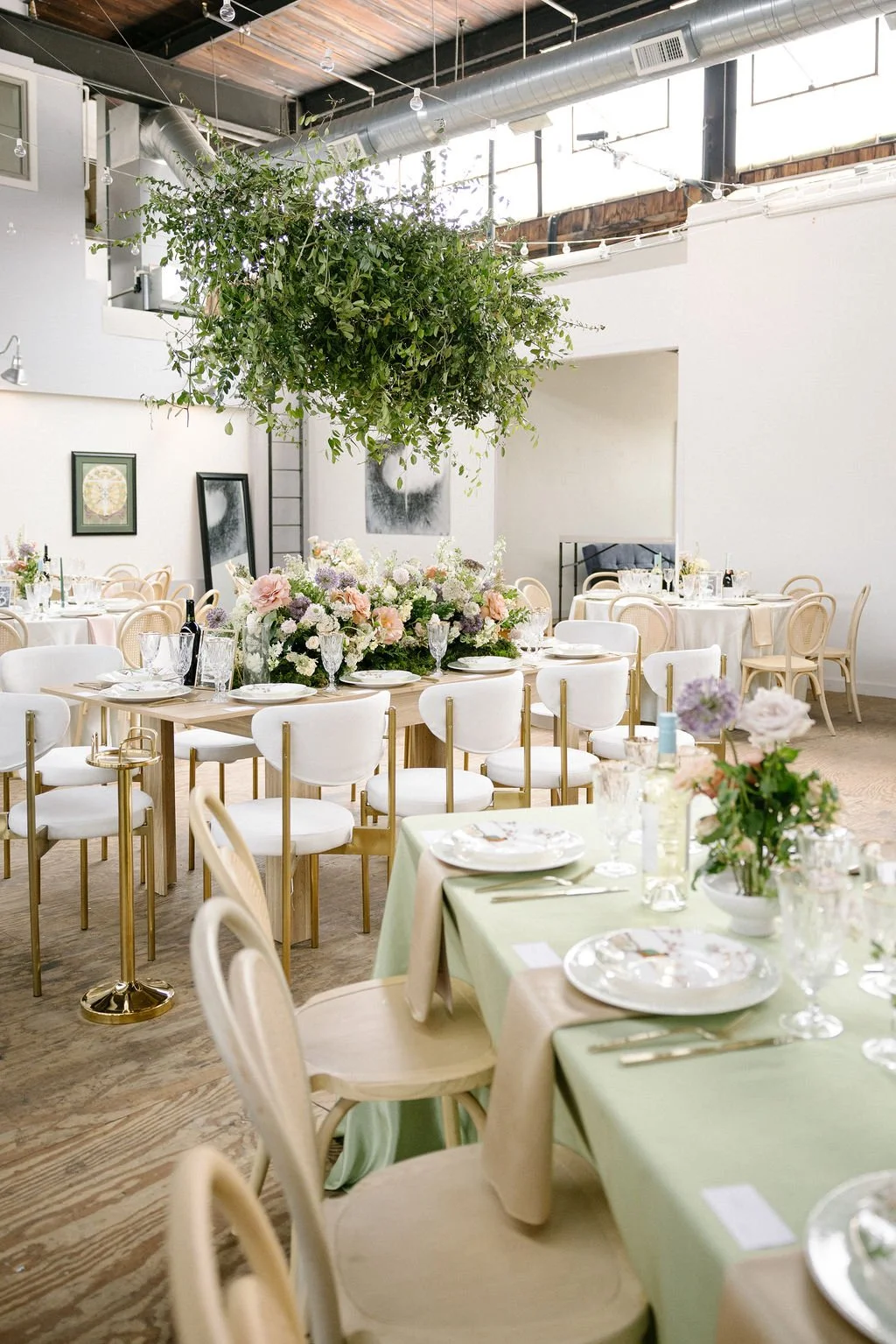 Elegant indoor event space decorated with floral arrangements and round tables set with white plates, glassware, and cutlery, featuring a large hanging greenery installation.