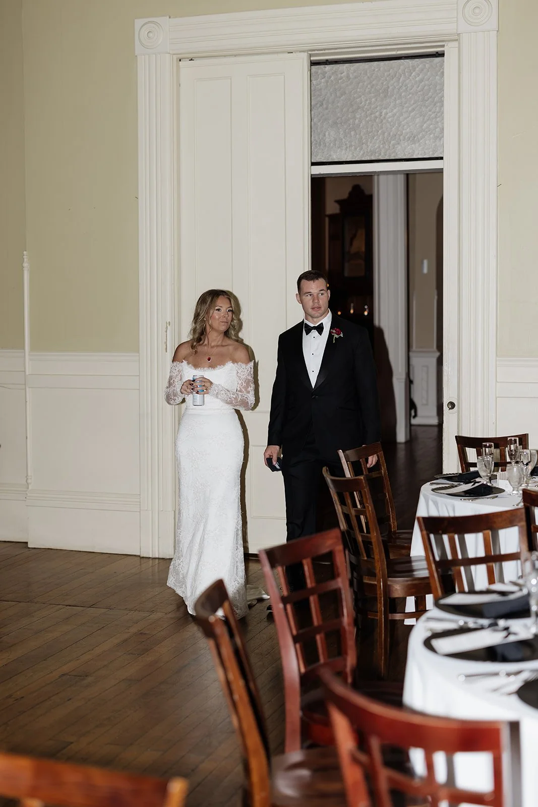 A bride in a long white gown and a groom in a black tuxedo walking through a doorway during a wedding reception.