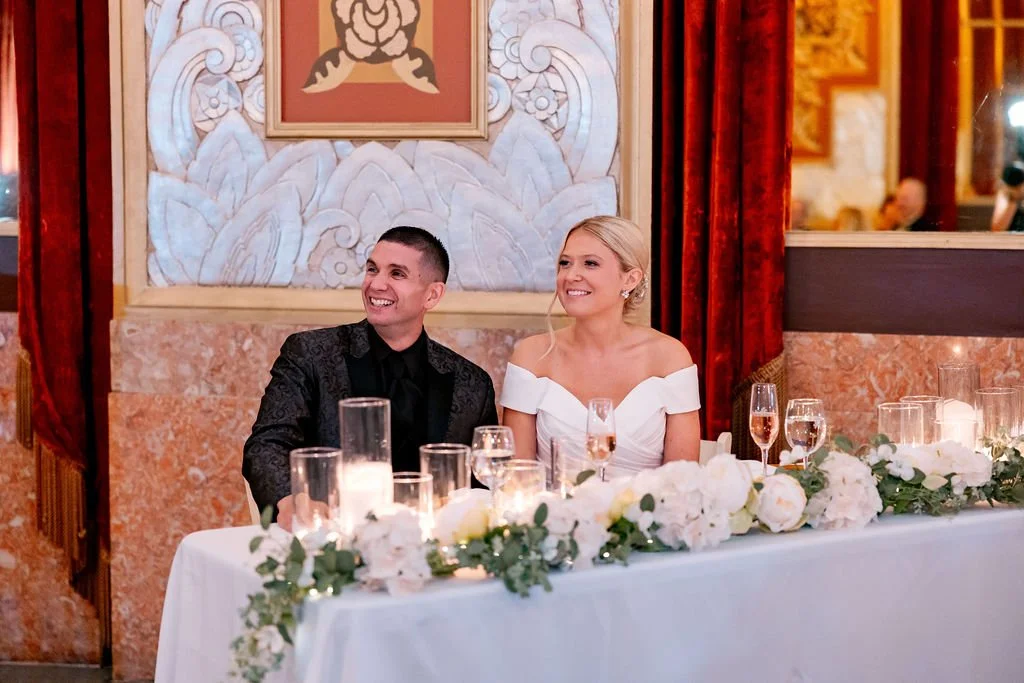 Bride and groom sitting at a decorated wedding reception table with flowers and candles, smiling and looking to the side in an elegant room.