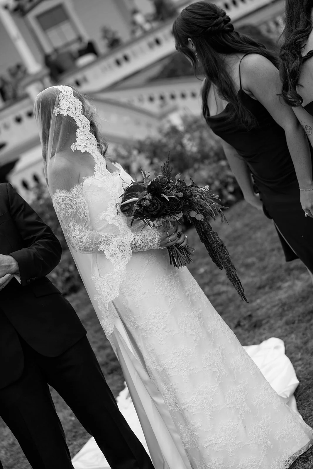 A black-and-white photo of a bride in a lace wedding dress holding a bouquet of flowers, standing outdoors with other guests, two women, nearby.