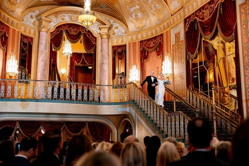 A wedding ceremony taking place on a grand staircase inside an ornate, luxurious venue with gold and red decor, chandeliers, and tall windows with red curtains. A bride in a white dress holding a bouquet and a groom in a tuxedo are descending the sta