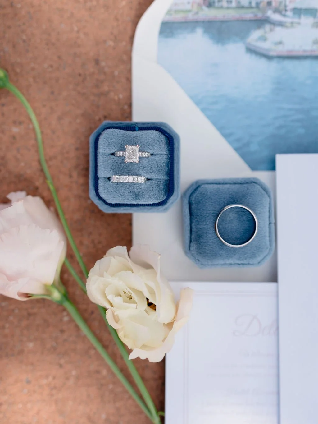 Diamond engagement ring and wedding band in blue velvet ring boxes, with white flowers and a postcard in the background.