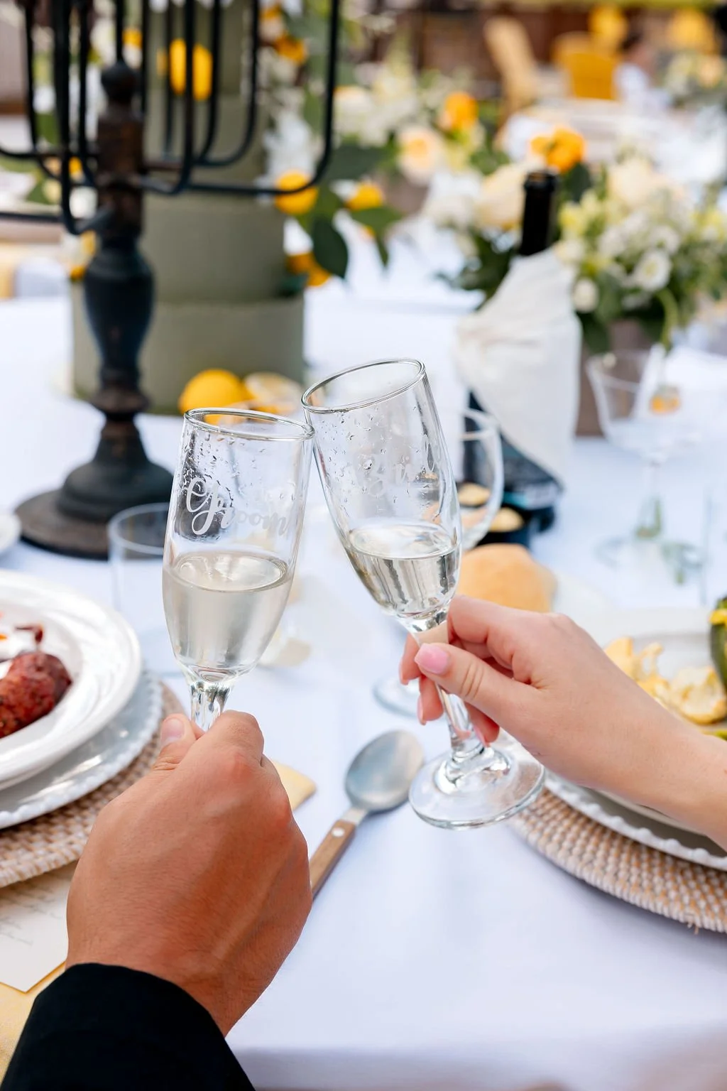 Two hands holding champagne flutes toasting at an indoor or outdoor gathering with a decorated table, flowers, and a candelabra in the background.