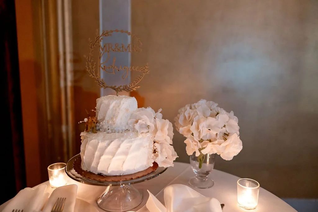 Wedding cake with white flowers and a topper that says "Mr & Mrs" with names and a date, surrounded by candles and flowers on a table.