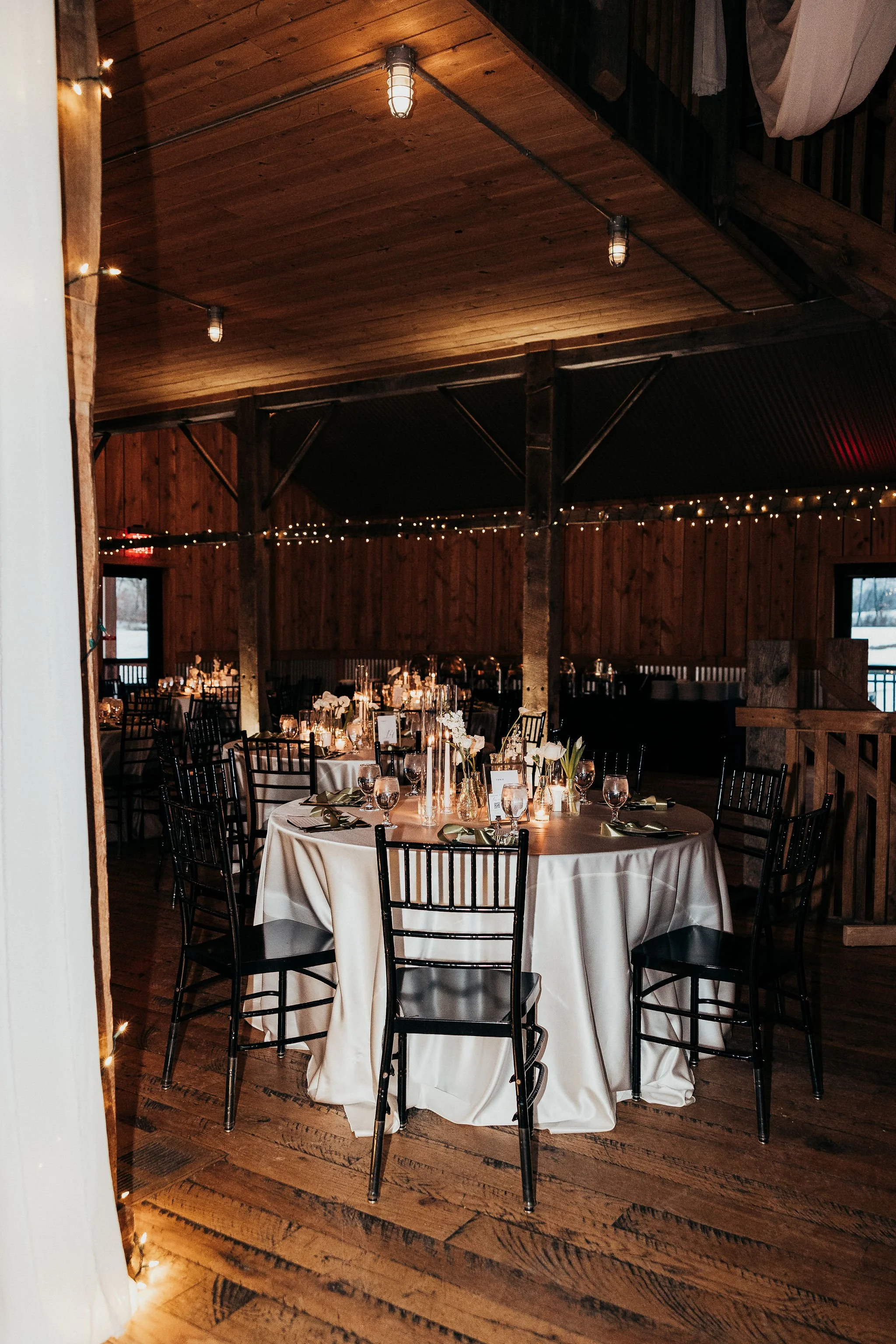 Wedding reception or event setup with round tables covered in white tablecloths, elegant table settings, and black chairs. The room has wooden walls and ceiling decorated with string lights, with warm ambient lighting.