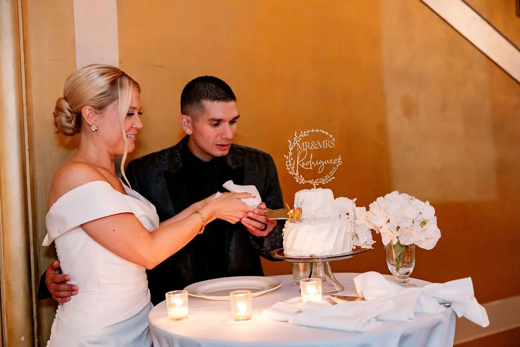 A bride and groom cut a wedding cake together at their reception, with candles and flowers on the table.