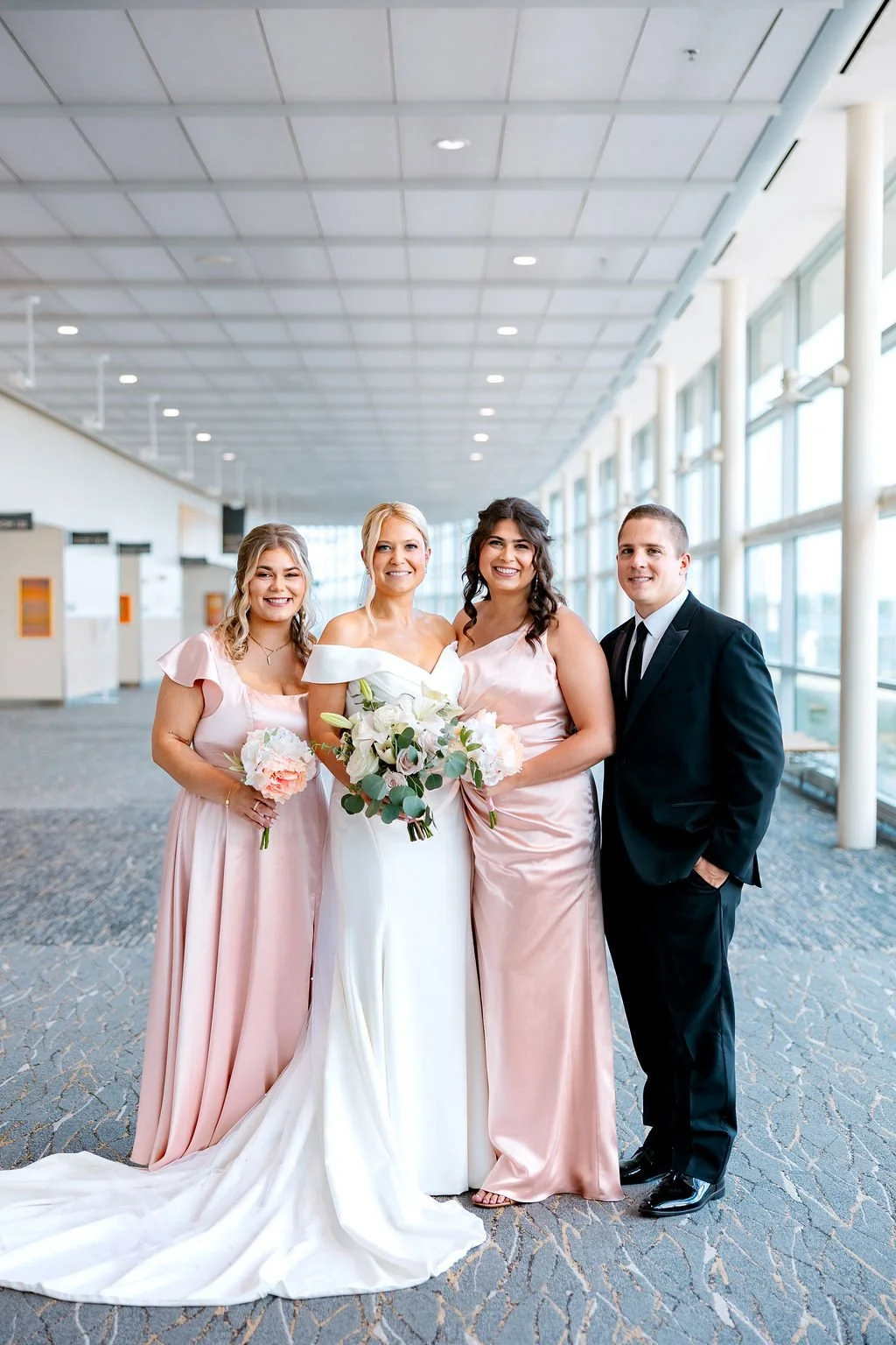 A bride and wedding party standing in an indoor venue with large windows. The bride wears a white gown and holds a bouquet of white flowers and greenery, flanked by two bridesmaids in blush pink dresses holding smaller bouquets, and a groom in a blac