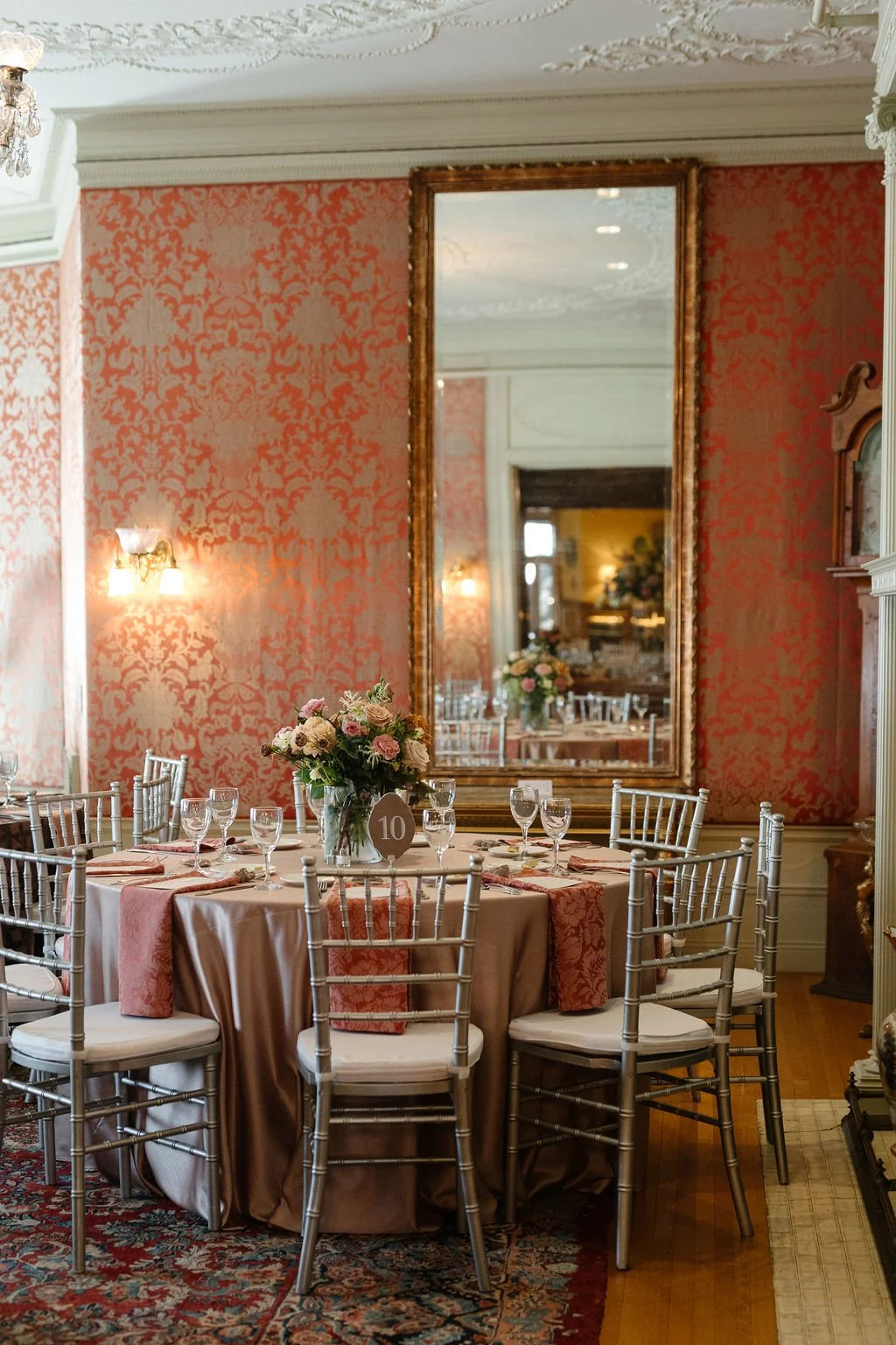 Elegant dining room decorated for a special event with a round table, silver chairs, a floral centerpiece, tableware, and a large mirror on a pink patterned wall.