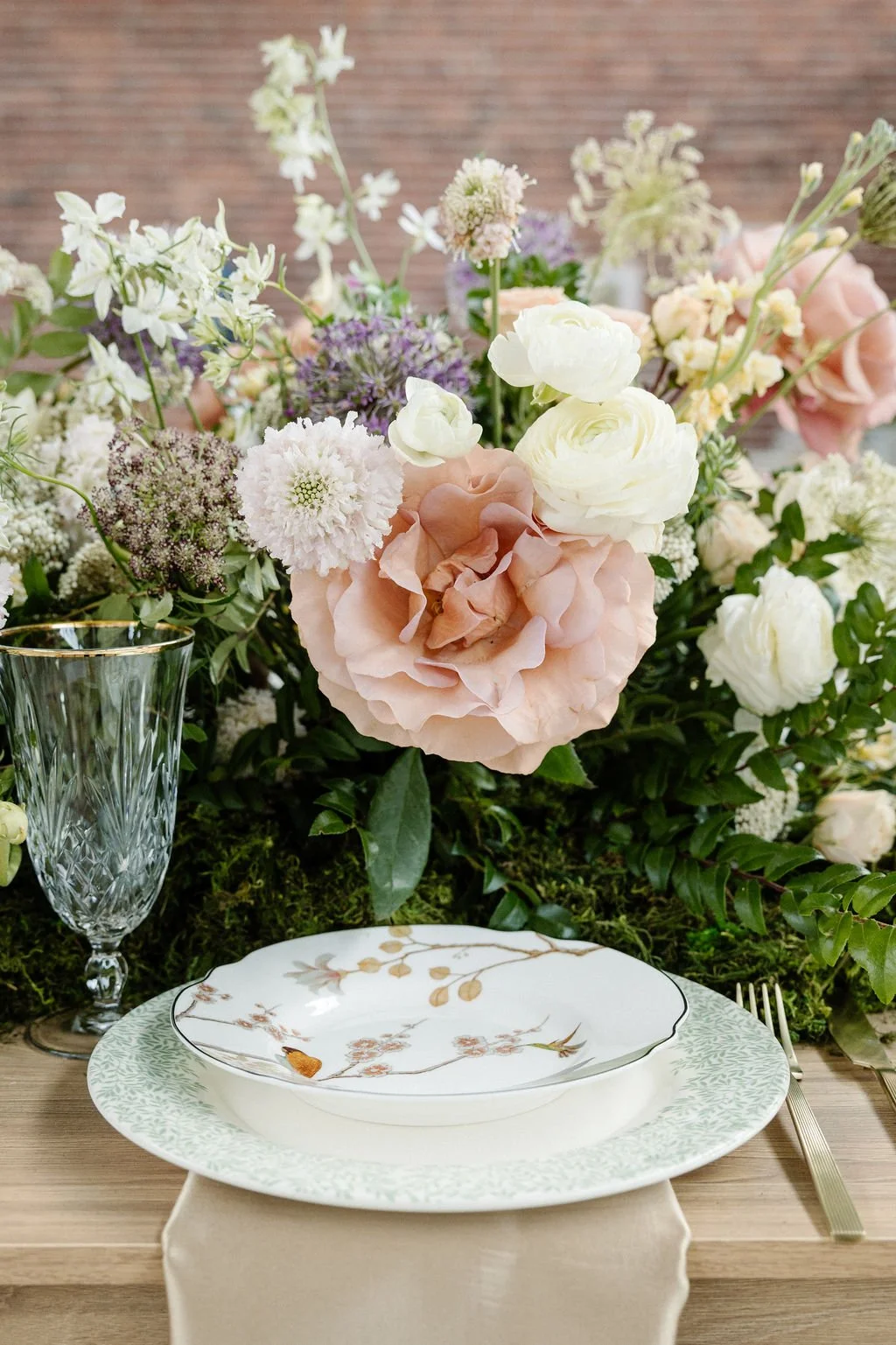 Elegant table setting with fine china, a crystal glass, and a floral centerpiece of pink and white flowers, including roses and other blooms.