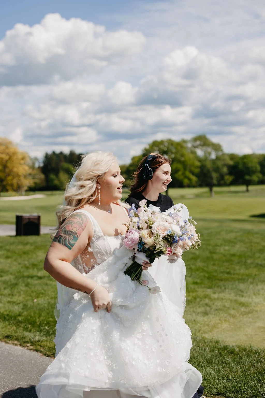 Two women, one in a wedding dress holding a bouquet, walking outdoors in a park with green grass and trees, under a partly cloudy sky.