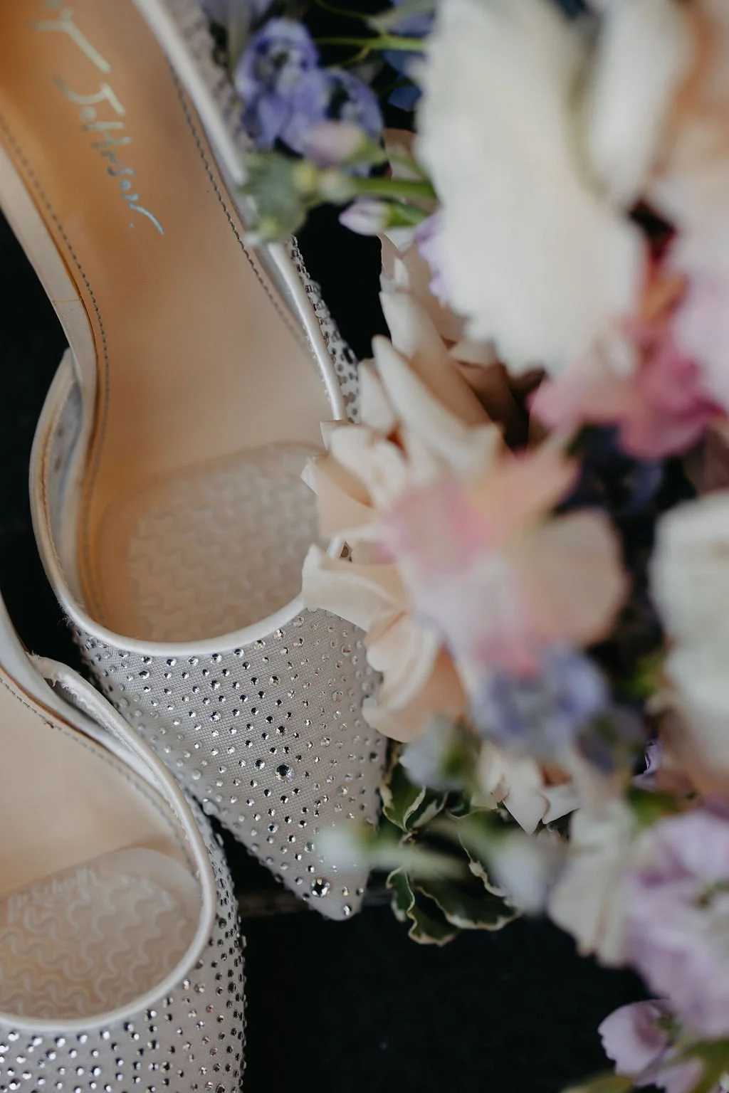 A pair of white high-heeled shoes embellished with small rhinestones and a flower arrangement with pink, white, purple, and cream-colored flowers.