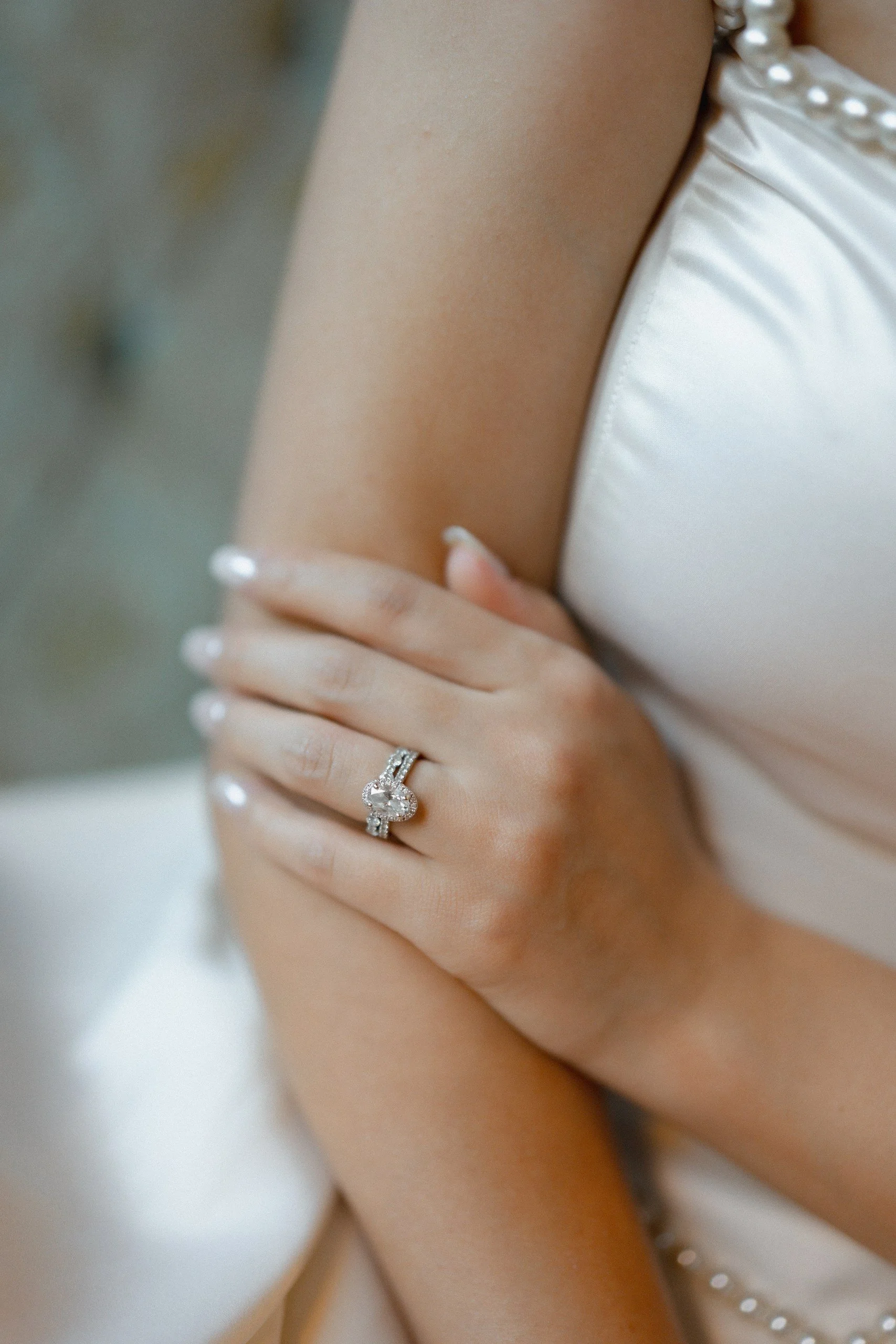Close-up of a woman wearing a ring with a large, clear gemstone on her finger, and pearl jewelry on her white satin dress.