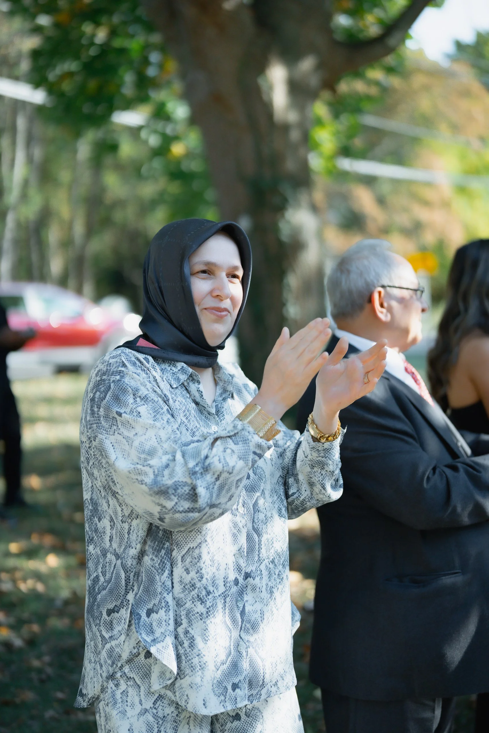 A woman in a snake-print shirt and matching pants, wearing a black headscarf and gold jewelry, clapping at an outdoor gathering during daytime with other people and trees in the background.