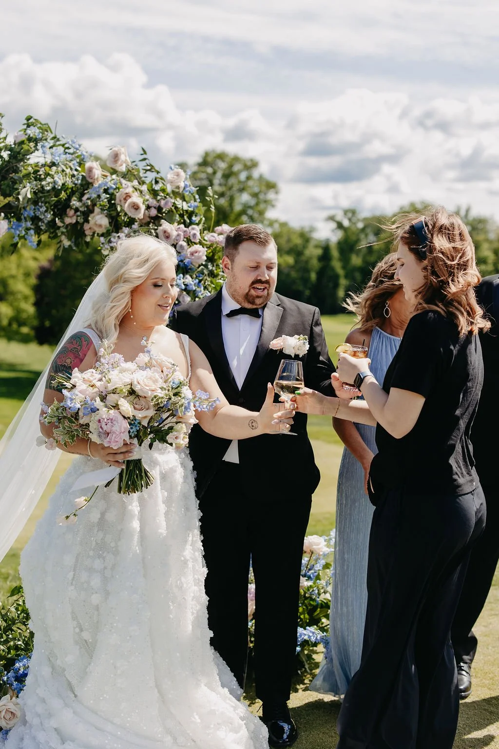 A wedding ceremony outdoors with four people, including a bride with blonde hair in a white dress holding a bouquet, a groom in a tuxedo with a boutonniere, and two women dressed in blue and black, celebrating with drinks under a floral arch on green