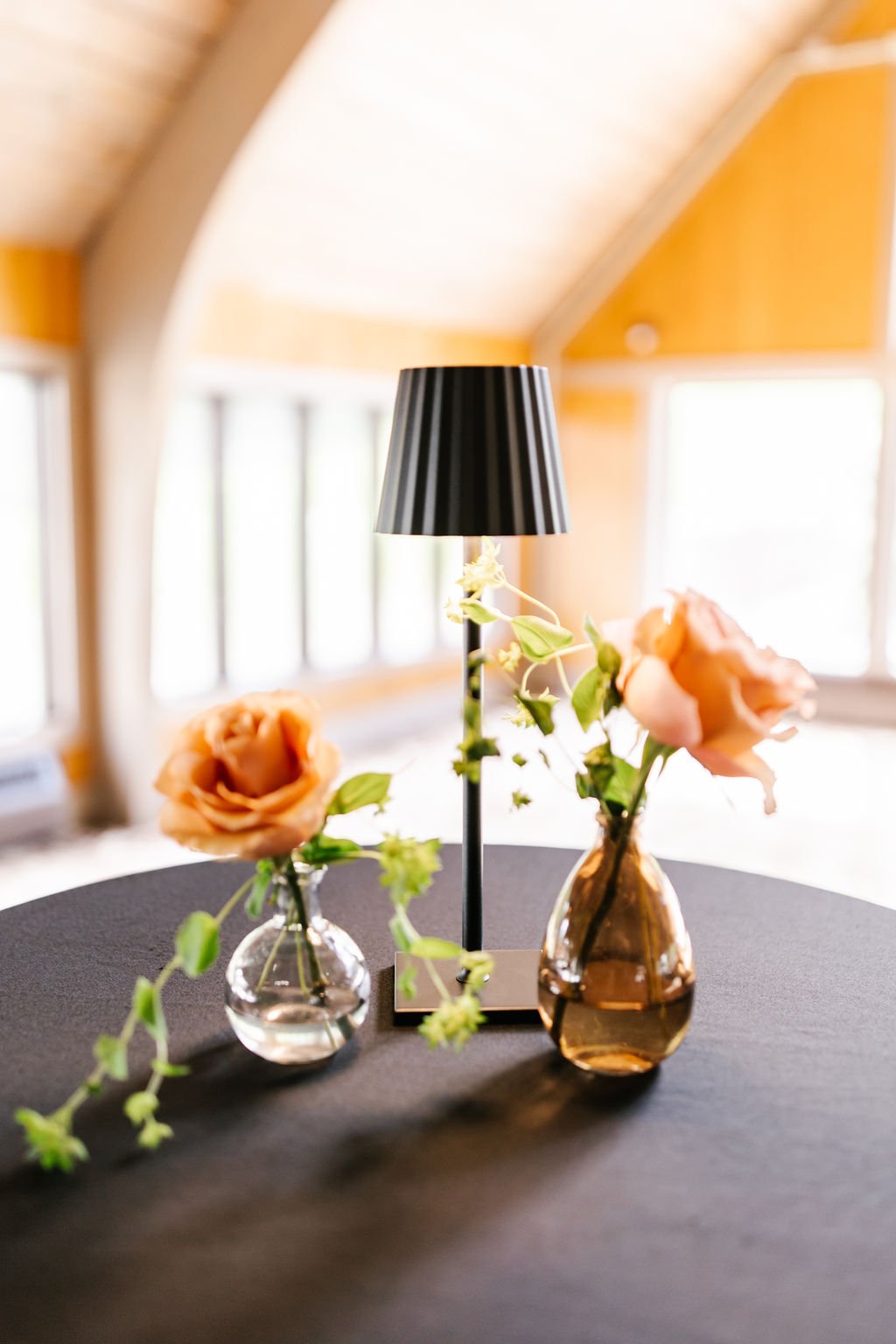 A small table decorated with two vases of roses, a black and gold lamp, and leafy green plants, set in a sunny room with windows and wooden paneling.