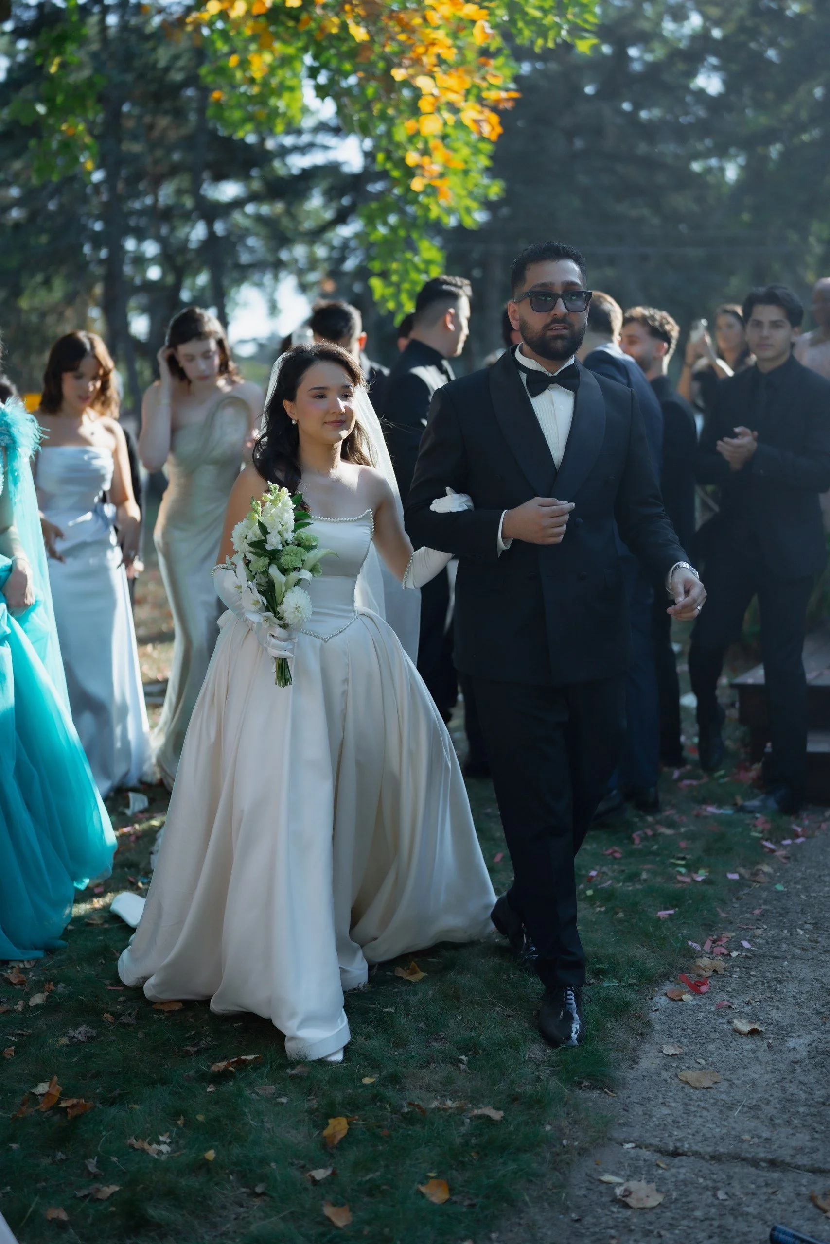 A group of people dressed formally for a wedding, walking outdoors on a sunny day with trees and fallen leaves in the background. The focus is on a woman in a gown holding a bouquet and a man in a tuxedo with sunglasses.