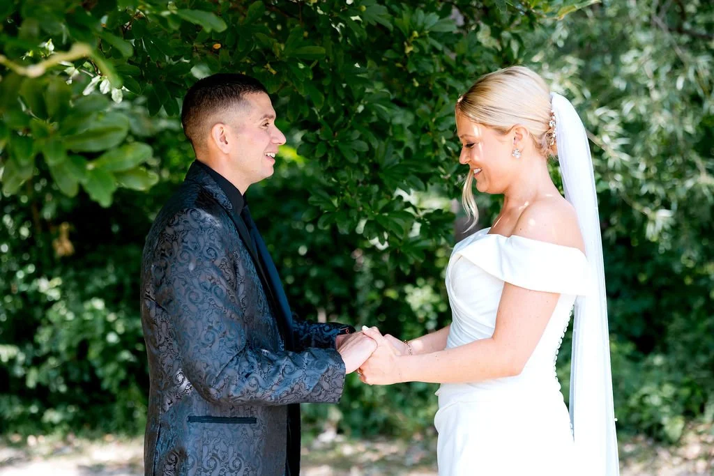 A bride and groom holding hands and smiling at each other outdoors surrounded by green foliage.