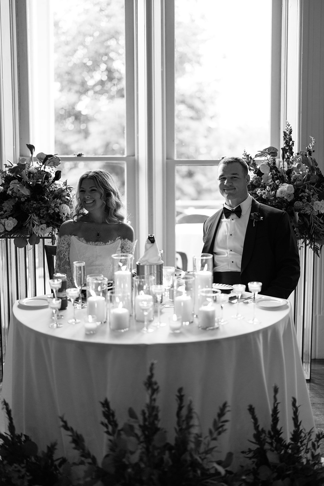 Black and white photo of a bride and groom sitting at a decorated table with candles and flowers, in front of large windows.
