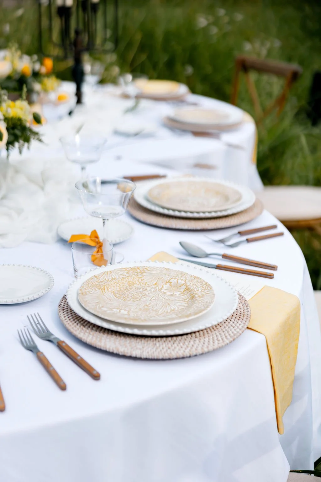Elegant outdoor dinner table set with white tablecloth, patterned plates, silverware, glassware, and floral centerpiece, surrounded by greenery.
