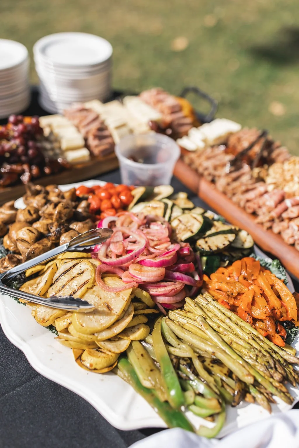 A platter of grilled vegetables including zucchini, asparagus, onions, and peppers, with a side of cherry tomatoes and other grilled vegetables, set on a black table with other dishes and empty bowls in the background.