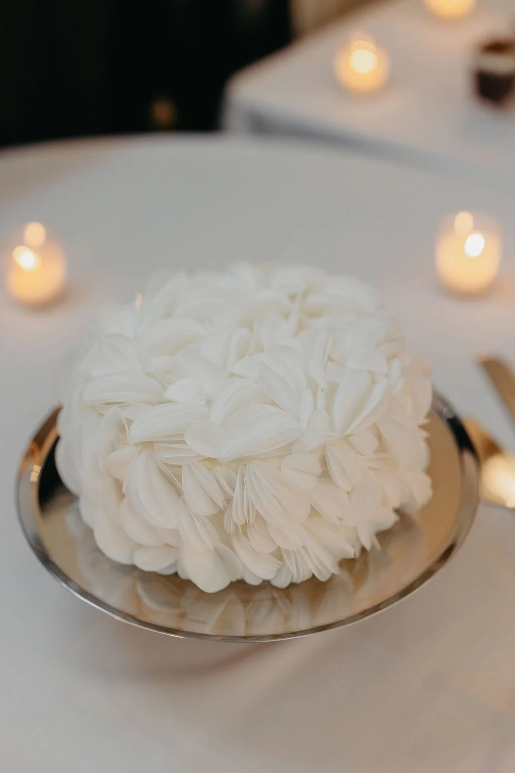 White floral arrangement on a silver tray, surrounded by small candles on a white table.