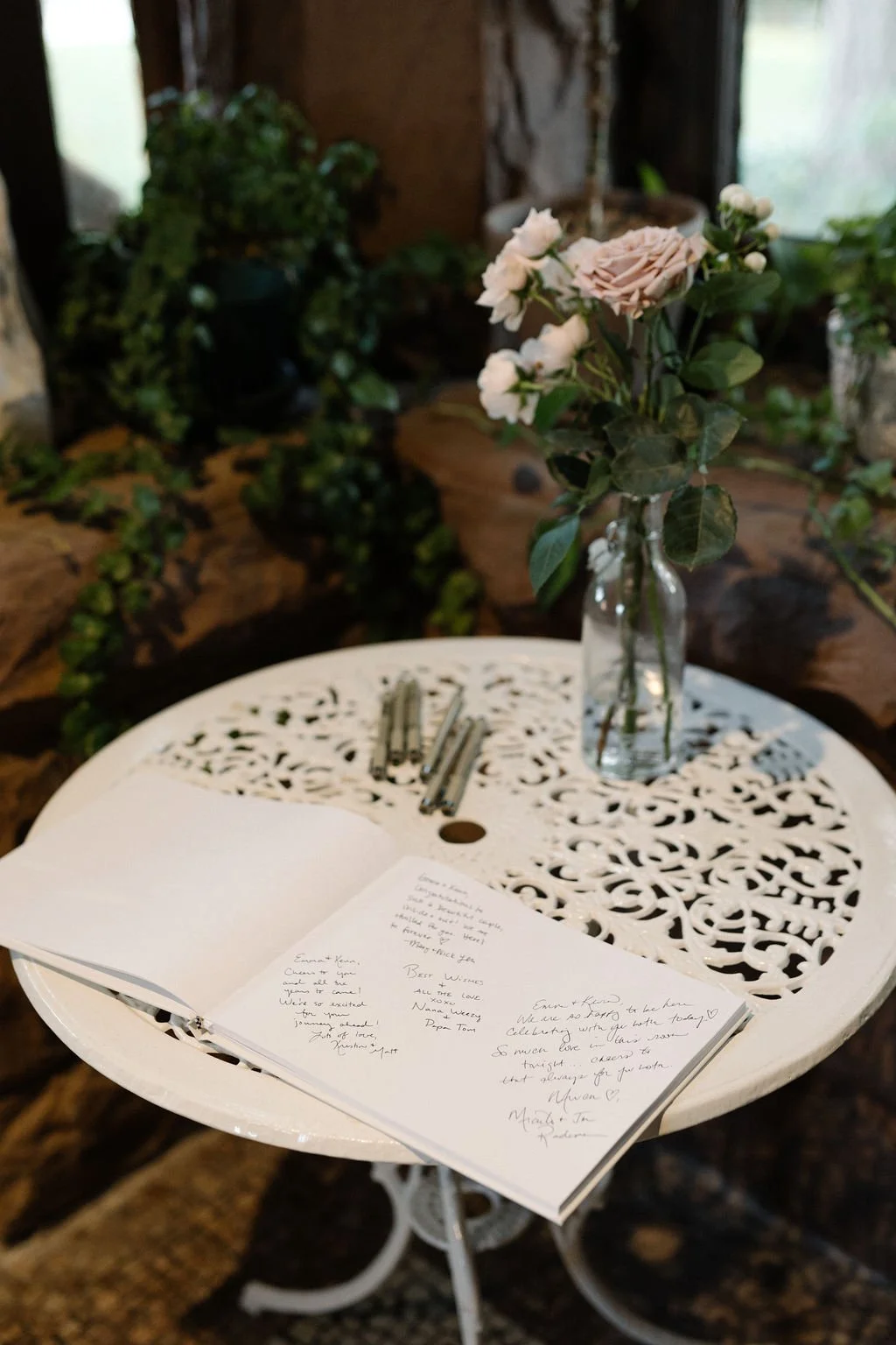 A white, ornate, circular table holding an open notebook, several silver pens, and a clear glass vase with pink and white roses. The background features greenery and a rustic wooden interior with a window.
